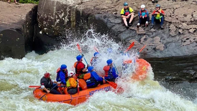 A group of people wearing helmets and life jackets paddle a whitewater raft through a strong current on the Gauley River in West Virginia. In the background, another group of people sits on a large rock, watching the raft navigate the rapids. Photo by Matt Kearns, USGS 