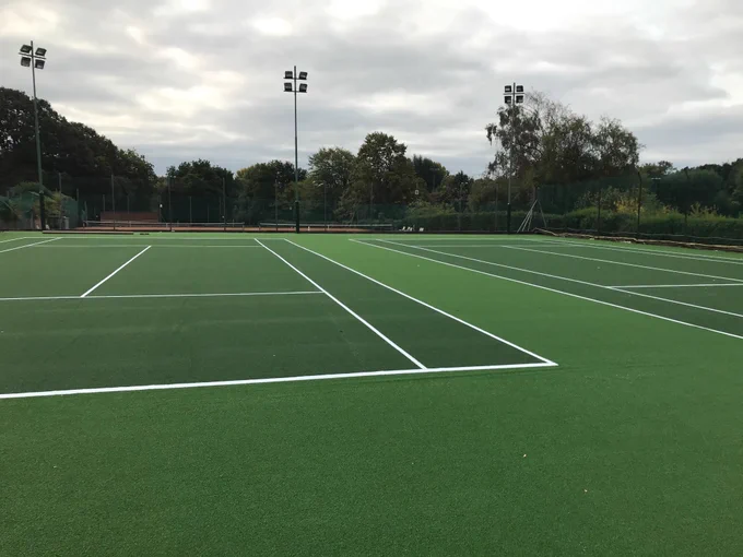 Empty outdoor tennis courts with green surfaces and white boundary lines under a cloudy sky.