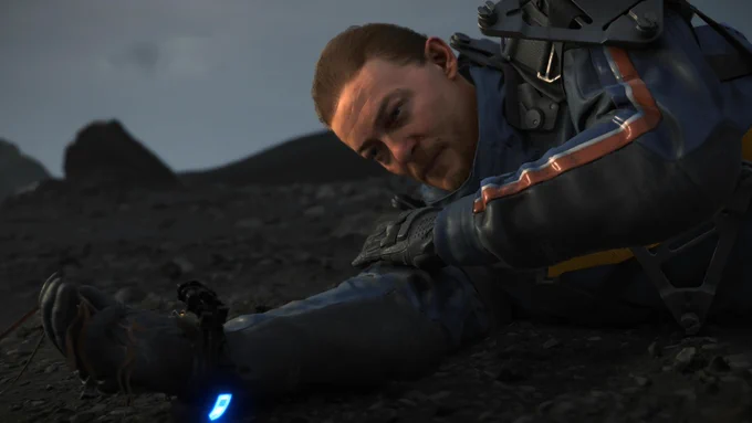 Eine Nahaufnahme von Sam Bridges der in Death Stranding Directors Cut auf einem felsigen Boden liegt. Er hat einen intensiven Gesichtsausdruck und Schweiß steht auf seiner Stirn. Ein Teil seines blauen Anzugs und seine Hand sind gut sichtbar.