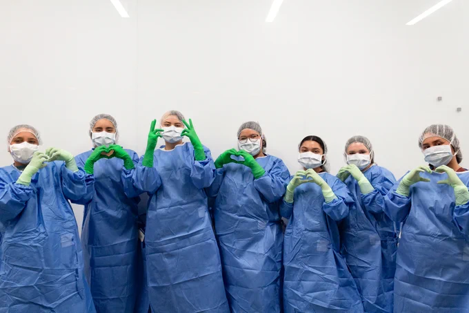 A group of healthcare professionals in blue surgical gowns, masks, and hairnets stand in a line, forming heart shapes with their hands. They wear green gloves and are in a medical environment with a white background.