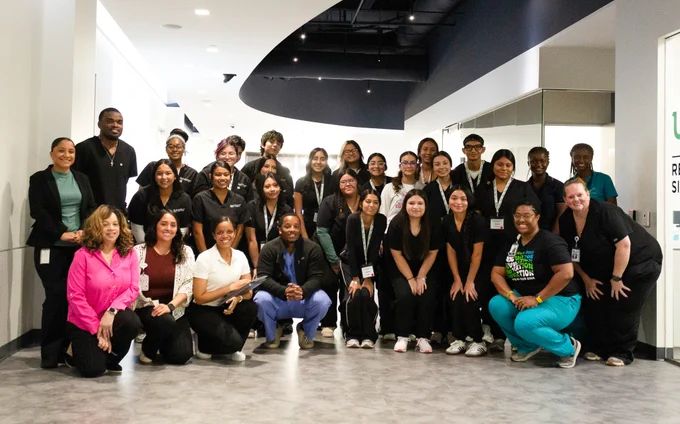 A group of people, dressed in a mix of business and casual attire, are posing together inside a modern building. Some wear lanyards, suggesting they may be part of an event or team. There is a sign in the background with partially visible text.