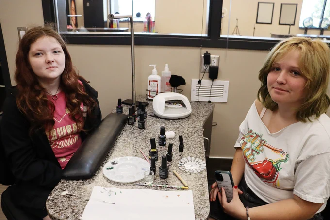 Two women sit at a nail salon table with nail polish bottles, a UV lamp, and manicure tools arranged on the countertop.
