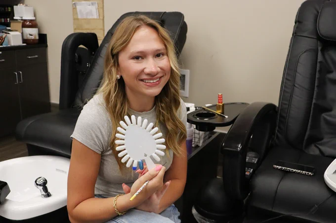 Woman holding a nail polish color wheel and brush, sitting in a salon chair next to a pedicure station.