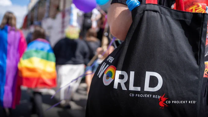 A black Open World tote bag with a rainbow eye logo hangs from a participant’s shoulder during a Pride parade. People, rainbow flags, and colorful balloons blur in the background.