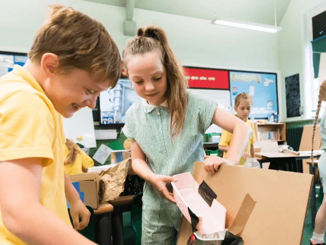 Two students in a classroom are smiling while working together on a craft project using cardboard and tape. 