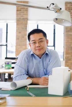 Asian man wearing glasses and a light blue collared shirt, sitting at a desk with some papers.
