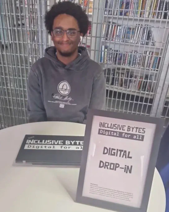 A young person sat at a table with a sign and a laptop. The sign reads "Inclusive Bytes Digital For All. Digital Drop-In"