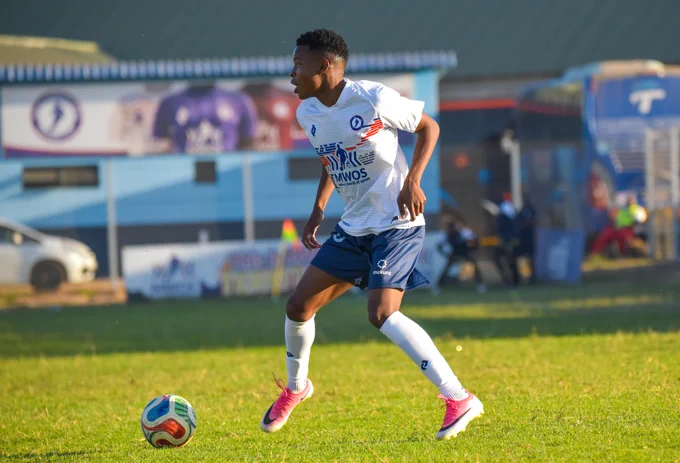 A soccer player in a white and blue uniform is in motion during a game on a grassy field. The player is focused on the ball, with a vibrant sports atmosphere in the background.