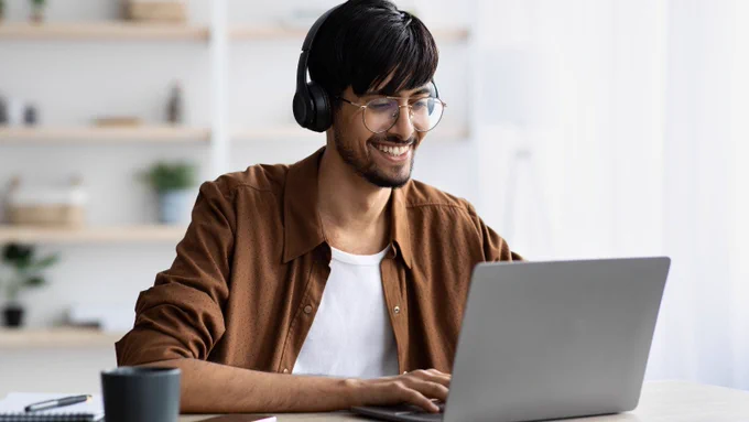 A person sat a desk, wearing headphones and smiling, as they work on a laptop.