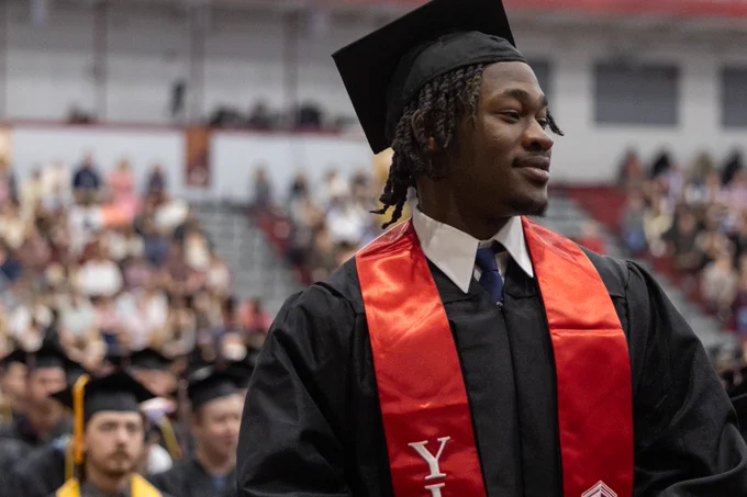Graduate wearing a black cap and gown with a red stole standing in a crowded auditorium during a graduation ceremony.
