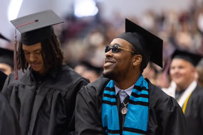 Two graduates in black caps and gowns sit among a crowd during a graduation ceremony, one wearing a blue and black stole.