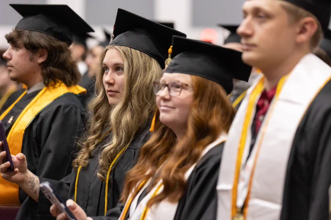 Group of graduates in black caps and gowns with gold stoles standing in a row during a ceremony.