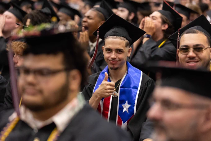 Graduate wearing a cap and gown with a stole giving a thumbs-up during a commencement ceremony.