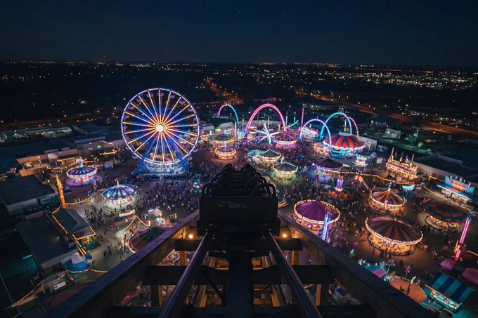 At the highest point of a roller coaster at night, looking down at all the colorful lights from the fairgrounds