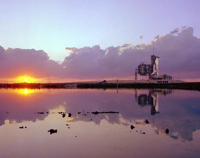 On the right, Space Shuttle Columbia is lit by spotlights at Launch Pad A at NASA's Kennedy Space Center while on the left, the sun dips below the horizon in this photo taken at sunset on February 19, 1981. The scene, dominated by lavender clouds looming along the horizon, is reflected in a lagoon in the foreground.
