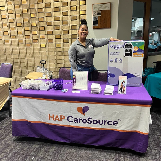 A person stands behind a table covered with a purple and white tablecloth featuring the HAP CareSource logo. On the table are brochures, and informational materials. The background showcases a wall with decorative blocks.