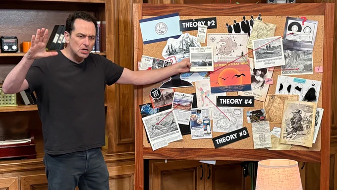 Matthew Rhys gestures emphatically at a cork board filled with pinned theories, newspaper clippings, and other clues.