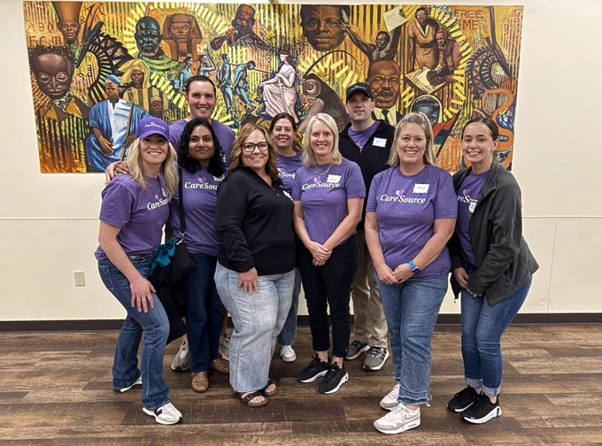 A group of nine people standing indoors, smiling. Many wear purple "CareSource" shirts. A colorful mural depicting various figures is on the wall behind them.