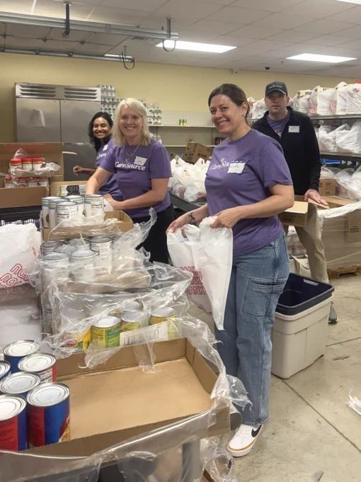 Volunteers are organizing food items in a pantry. They are sorting canned goods and packing them into bags on a table. The setting appears to be a storage room with shelves stocked with various supplies. A person in the back is smiling and observing.