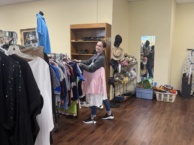 A person is browsing through clothing racks in a thrift store. The room has wooden floors and shelves with shoes and accessories. A large mirror reflects various items, including hats. The atmosphere is casual and inviting.