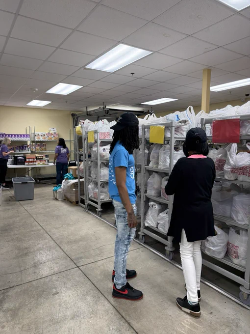 People are standing in a room filled with shelves holding bags of groceries. The space resembles a food pantry or distribution center, with organized items and visible labels on bags.