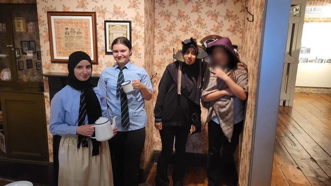 A group of students wearing Victorian-themed outfits and school uniforms, posing indoors. The setting includes floral wallpaper and framed pictures suggesting a historical or educational venue.