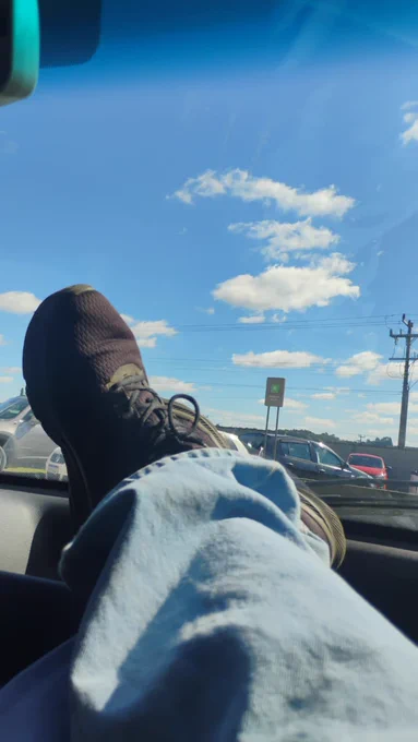 Foto vertical tirada do ponto de vista do passageiro dentro de um veículo. Uma perna vestindo calça jeans azul-clara está relaxada, com o pé calçando um tênis esportivo escuro apoiado sobre o painel do carro. Através do vidro do para-brisa, destaca-se um céu azul intenso salpicado de nuvens brancas e fofas. Ao fundo, um estacionamento ao ar livre com alguns carros e um poste de energia sob a luz do dia.