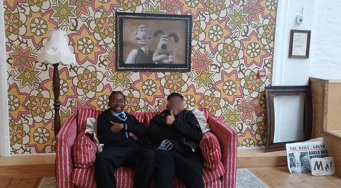 Two individuals sit on a red-striped sofa in front of a vibrant retro-patterned wall with a cartoon artwork of two characters. The atmosphere is casual and fun.