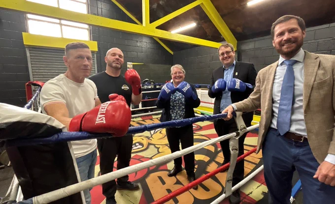 Five people in boxing gloves stand in a boxing ring, some in casual wear, others in suits.