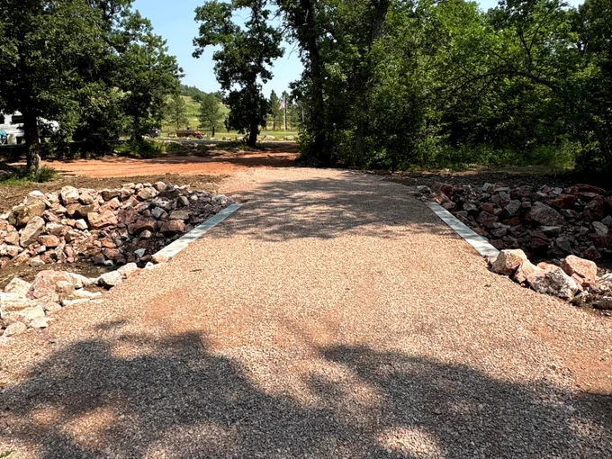 A newly constructed gravel pathway crossing a small rocky drainage area surrounded by green trees on a sunny day.
