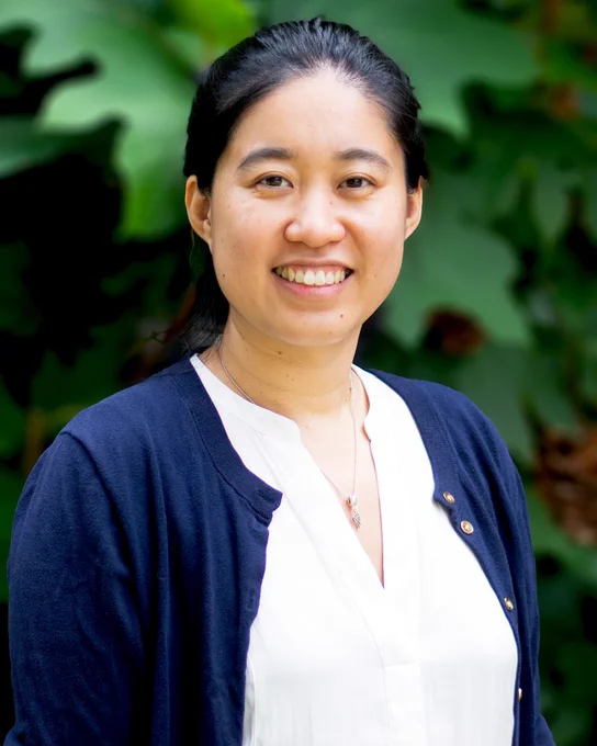 Woman wearing a white blouse and navy cardigan standing outdoors with green foliage in the background.