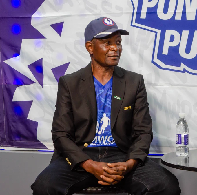 A man in a black blazer and cap sits at a table with a water bottle. A branded banner is in the background, and he appears to be engaged in a conversation or interview.