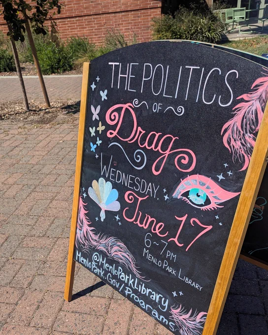 A standing chalkboard outside the library is colorfully decorated with drawings of feathers, butterflies, and an extravagantly made-up eye. Text: The Politics of Drag - Wednesday, June 17, 6-7 p.m., Menlo Park Library. @menloparklibrary www.menlopark.gov/programs