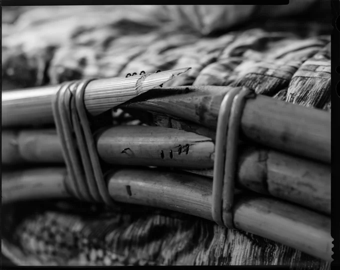 black and white large format picture showing three bent pieces of bamboo forming a bed's frame, held together by rattan, that have broken at the bend point and been stapled together. The top one has clearly broken through the staples