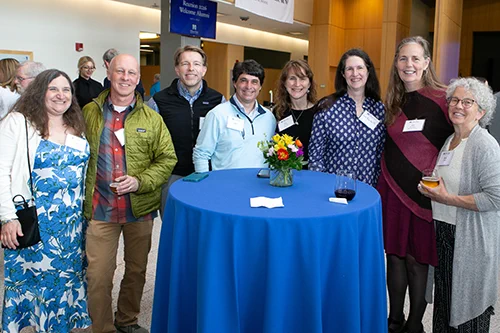Group of seven people standing around a tall round table covered with a blue tablecloth and a small flower arrangement in a bright indoor setting.
