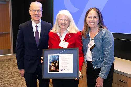 Three people stand together indoors, with the center person holding a framed certificate featuring a building image and text.