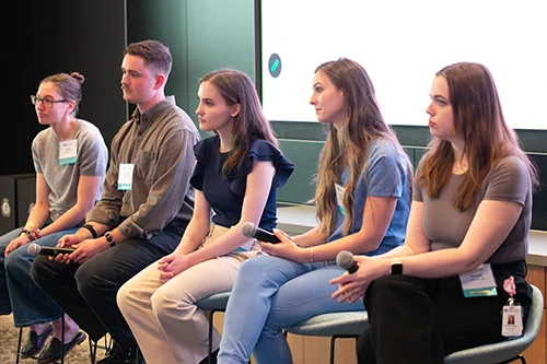 Five panelists seated in a row holding microphones during a discussion or Q&A session in a modern conference room.