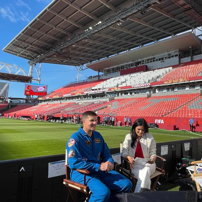 An astronaut in a blue flight suit speaking at a presentation.
An astronaut standing in front of a green screen during a media production.
An astronaut showing a garment to a group of children in a museum.
An astronaut in a blue flight suit being interviewed at a stadium.
