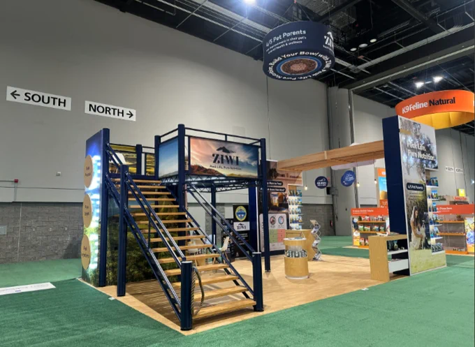 Custom double-deck trade show booth for Ziwi Pet Nutrition displayed at a convention center. The exhibit features a prominent staircase leading to a second-level meeting area, large branded graphics, wood-look flooring, product display shelving, overhead hanging signs, and open spaces for attendee interaction. The two-story design creates a strong visual presence while providing dedicated areas for meetings, product education, and brand engagement.