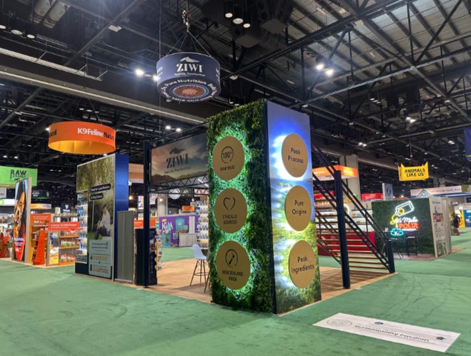 Corner view of the Ziwi Pet Nutrition double-deck trade show exhibit at a major industry event. The booth features illuminated graphic towers, a living green wall with brand messaging, large-format lifestyle imagery, an elevated second-story meeting space, and open product engagement areas. Overhead hanging signage enhances visibility, while the exhibit's natural textures and premium finishes reflect the brand's focus on quality ingredients and New Zealand sourcing.