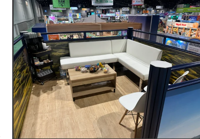 Private second-level meeting lounge inside the Ziwi Pet Nutrition double-deck trade show exhibit. The elevated space features comfortable white sectional seating, a wood-finish coffee table with refreshments, wood-look flooring, and panoramic views of the trade show floor below. Designed as a quiet hospitality area, the lounge provides a comfortable setting for customer meetings, relationship building, and business discussions away from the activity of the exhibit hall.