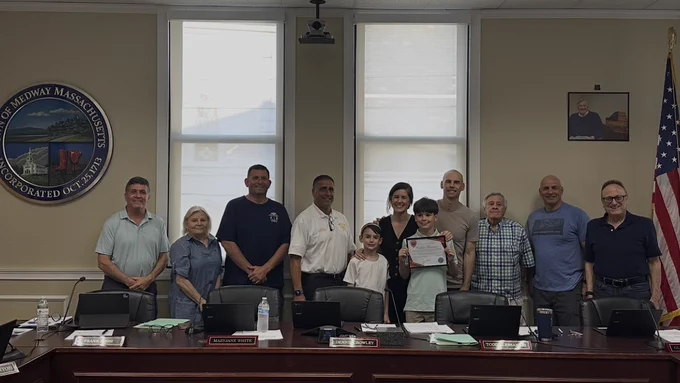 A group photo inside a municipal meeting room. A young boy standing in the center proudly holds an official certificate. He is surrounded by his family, members of the Select Board, and fire department leadership standing behind a long dark wood committee desk. On the wall to the left is the large circular Town of Medway, Massachusetts seal, and an American flag stands on the right.