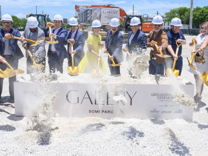 A group photo action shot of 12 adults wearing hard hats, shoveling dirt out of a large planter with the words "Gallery SoMi Parc"; as part of the ground breaking ceremony for the new building project.