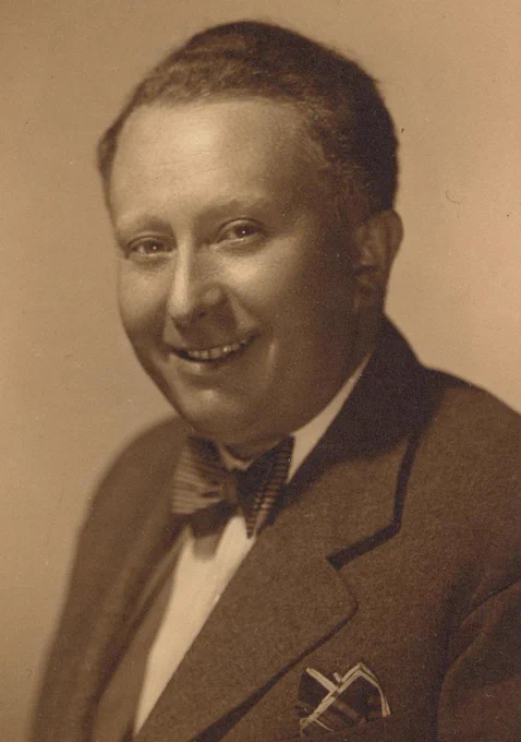 Sepia-toned portrait of a smiling man wearing a suit and a bow tie, featuring a pin on the lapel.
