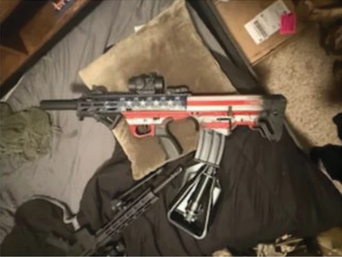 Semi-automatic rifle with an American flag design on the body, resting on a bed with pillows and blankets.