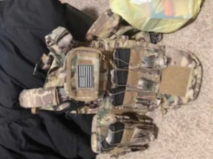 Camouflage tactical vest with multiple pouches and an American flag patch laid out on a carpeted floor.