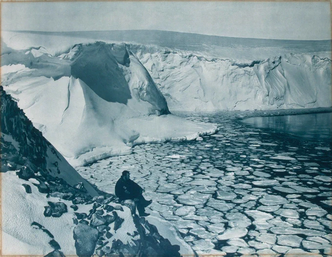 A photoprint of Commonwealth Bay, taken during the Australasian Antarctic Expedition in 1912 featuring an icy rocky cliff overlooking a bay with fractured ice chunks floating in the water.  A lone man sits on the edge of a cliff, looking out over the bay.