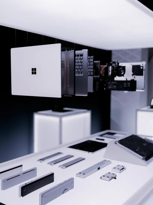 A display showcasing various components of a Microsoft device, including a white panel with the Microsoft logo, circuit boards, and metallic parts, arranged on a sleek white surface under soft lighting.