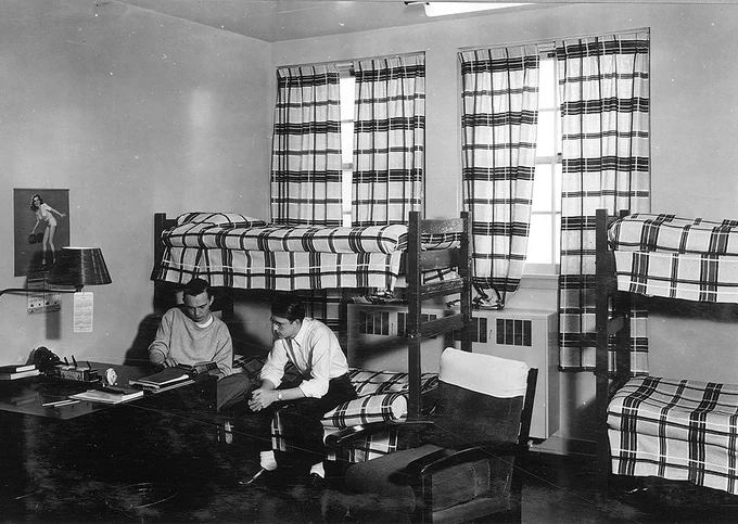 Two young men sit and talk in a dorm room with two sets of bunk beds and plaid curtains on the windows.