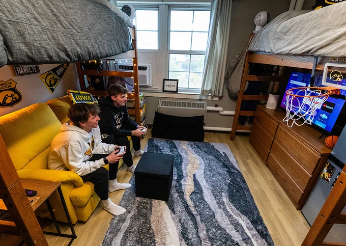 Two boys sitting on a yellow couch playing video games in a cozy dorm room with bunk beds and a basketball hoop on the dresser.